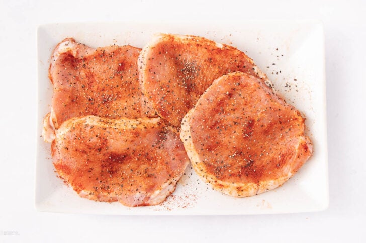 Four raw pork chops are arranged on a white rectangular plate, seasoned with red spices and black pepper, set against a white background.