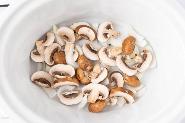 Sliced mushrooms and chopped onions in a white slow cooker, viewed from above.