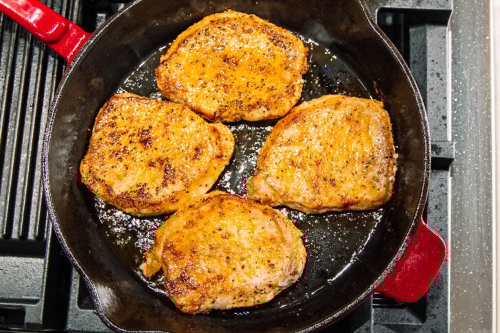 Four seasoned pork chops cooking in a black cast iron skillet on a stovetop, with a golden brown sear and some oil visible in the pan.