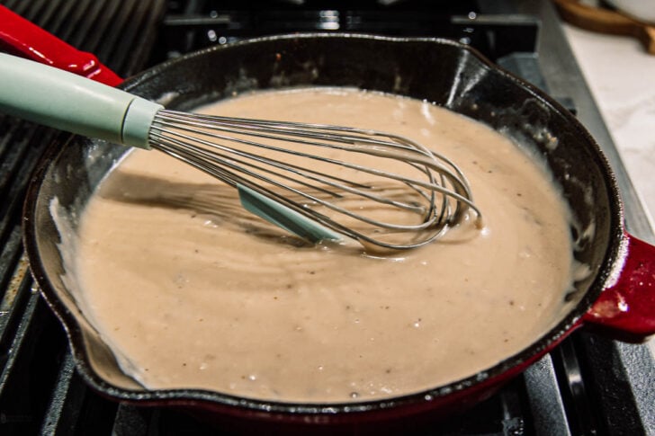 A close-up of a red skillet filled with creamy gravy being whisked on a stovetop. The metal whisk is mixing the thick, light brown sauce.