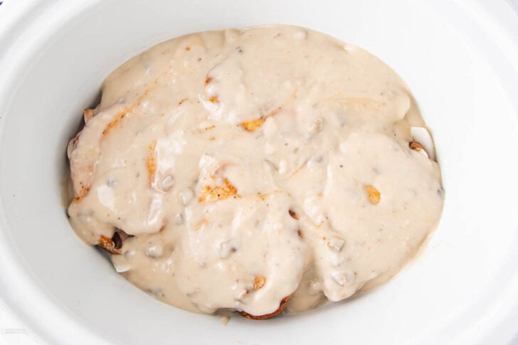 Chicken breasts covered in a creamy mushroom soup mixture inside a white slow cooker, ready to be cooked.