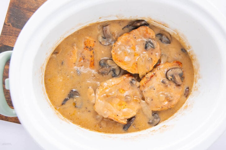 Three pieces of browned chicken in a creamy mushroom sauce inside a white slow cooker, with visible sliced mushrooms and onions in the sauce.