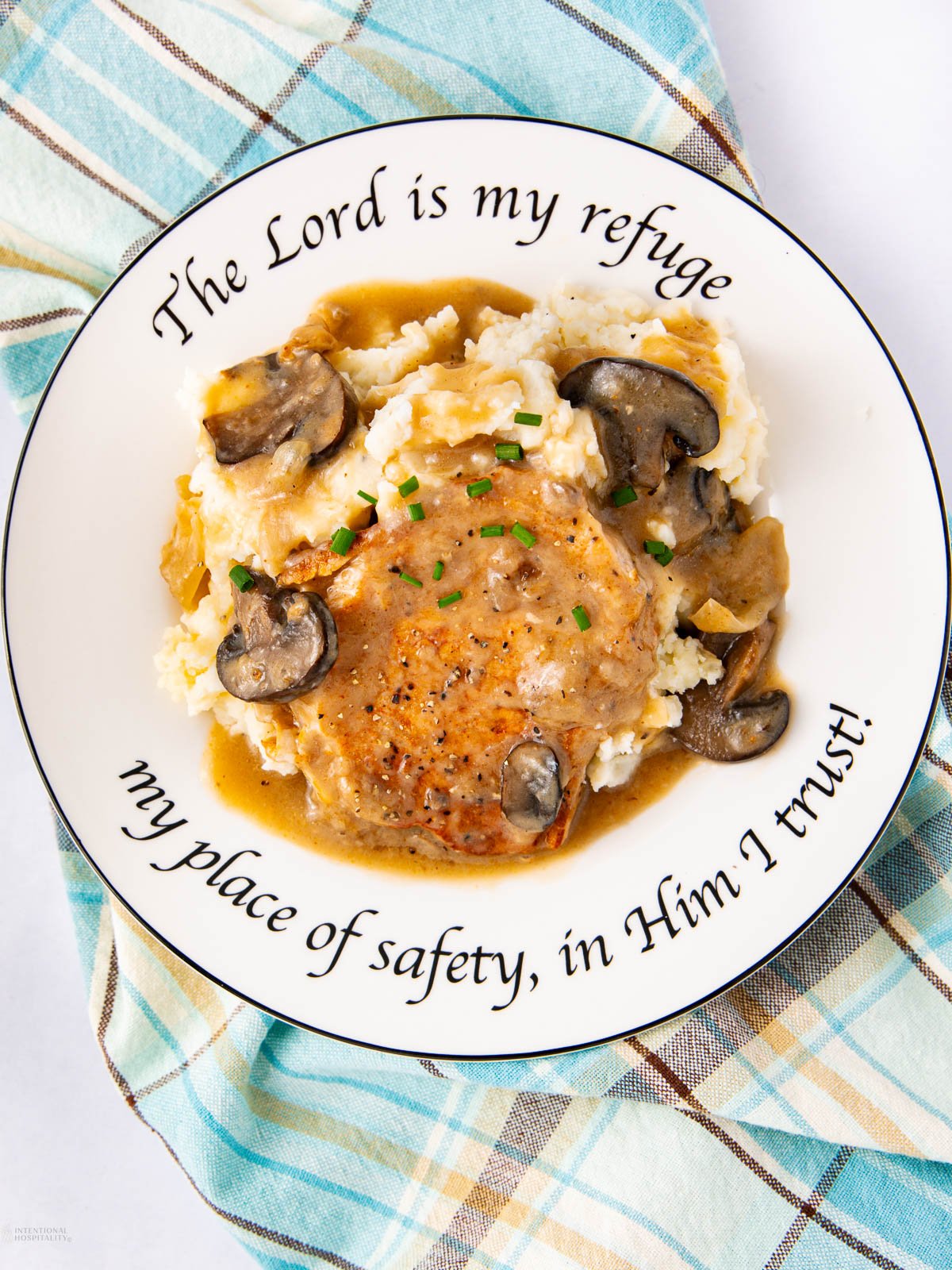 A plate of mashed potatoes topped with a piece of meat, creamy mushroom gravy, and chopped chives. The plate rim reads, "The Lord is my refuge, my place of safety, in Him I trust!" A plaid cloth is beneath the plate.