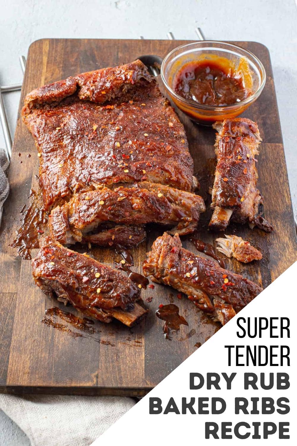A slab of baked ribs coated in dry rub sits on a wooden board, partially sliced into pieces, with a small bowl of barbecue sauce and a hint of canned cowboy candy recipe inspiration. Text overlay reads: "Super Tender Dry Rub Baked Ribs Recipe.