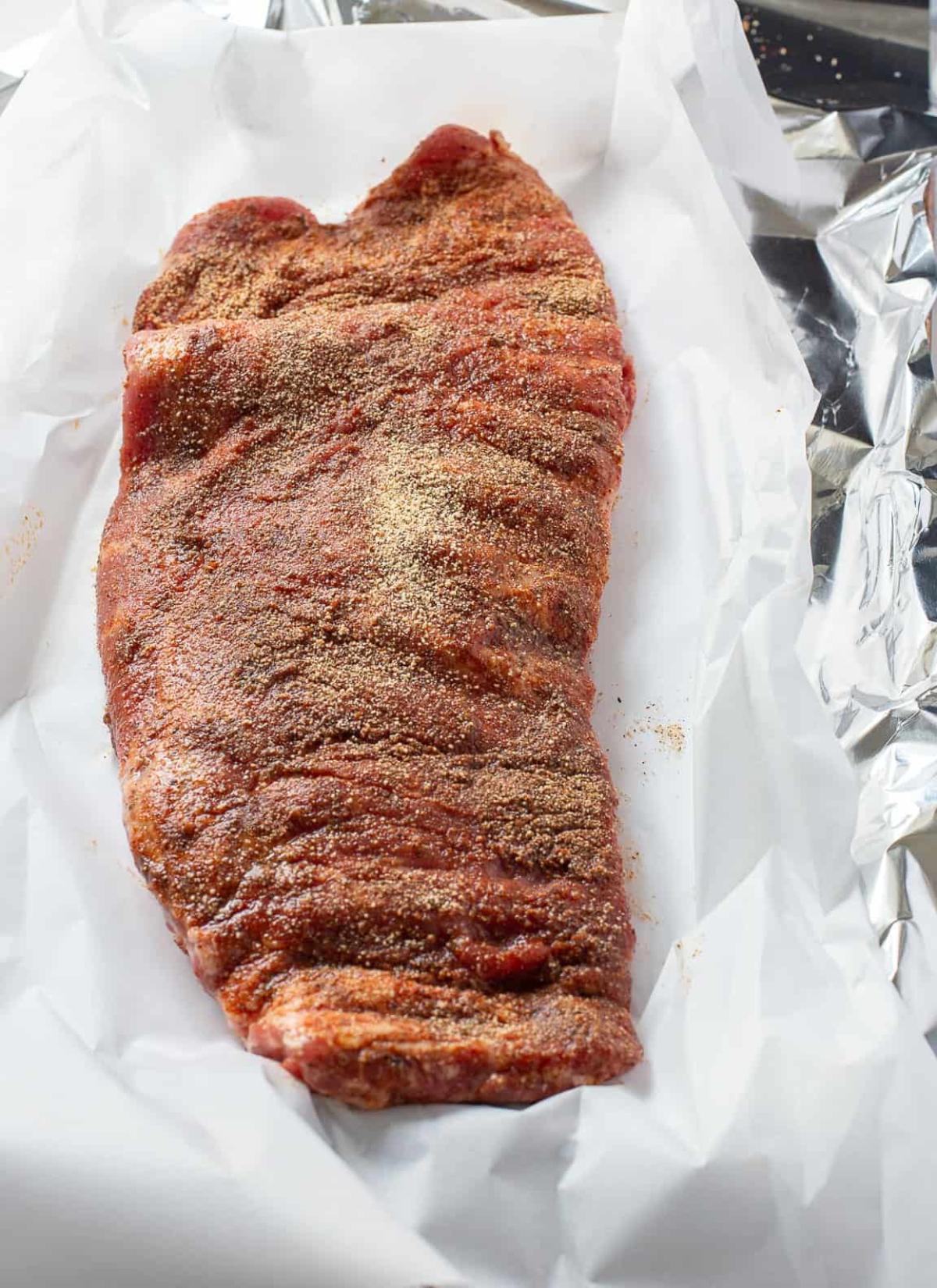 A slab of raw pork ribs coated with dry spice rub sits on white parchment paper, aluminum foil at the sides, ready to pair perfectly with a zesty canned cowboy candy recipe.