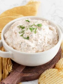 A white bowl filled with creamy dip seasoned with Thai spice blend and garnished with chopped herbs, placed on a wooden board next to ridged potato chips and a yellow cloth on a marble surface.
