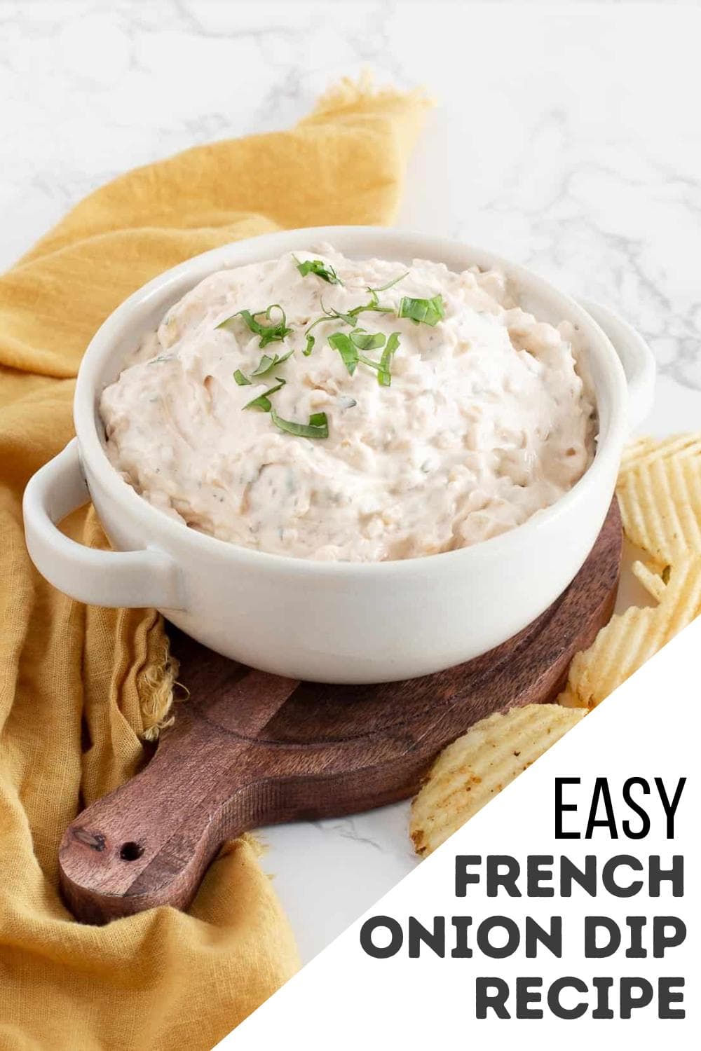 A white bowl filled with creamy French onion dip, enhanced with a hint of Thai spice blend and garnished with chopped herbs, sits on a wooden board next to wavy potato chips. A mustard-yellow cloth is underneath, with text reading "Easy French Onion Dip Recipe.