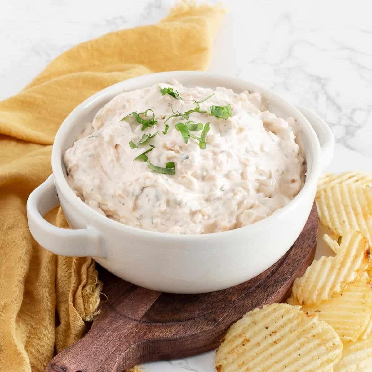 A white bowl filled with creamy dip seasoned with Thai spice blend and garnished with chopped herbs, placed on a wooden board next to ridged potato chips and a yellow cloth on a marble surface.