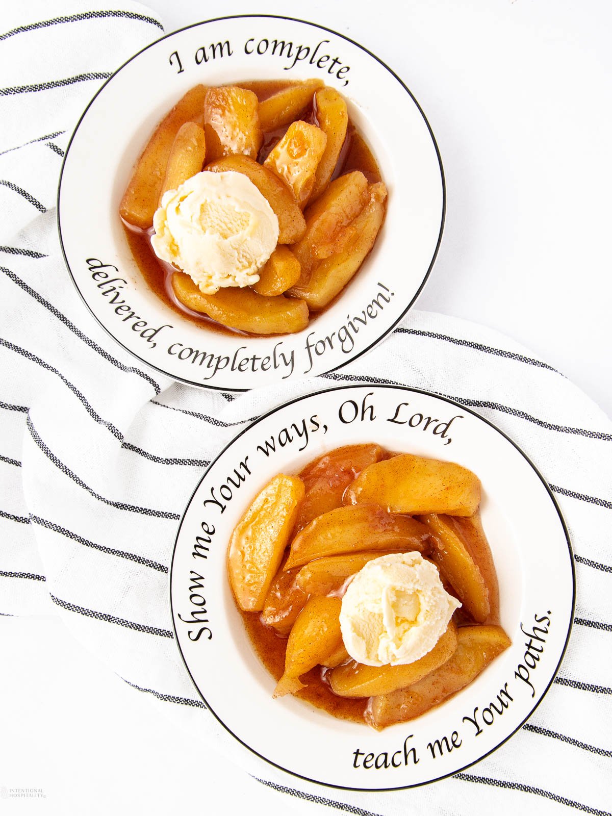 Two white bowls with black script hold baked apples in syrup, each topped with a scoop of vanilla ice cream. Inspired by a classic fried apples recipe, the bowls sit on a white surface with a striped cloth underneath.