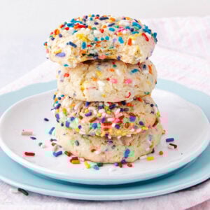 A stack of four sugar cookies covered in colorful rainbow sprinkles sits on a white plate with a blue plate underneath, with a light pink and white striped cloth in the background.