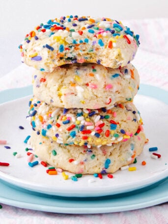 A stack of four sugar cookies covered in colorful rainbow sprinkles sits on a white plate with a blue plate underneath, with a light pink and white striped cloth in the background.