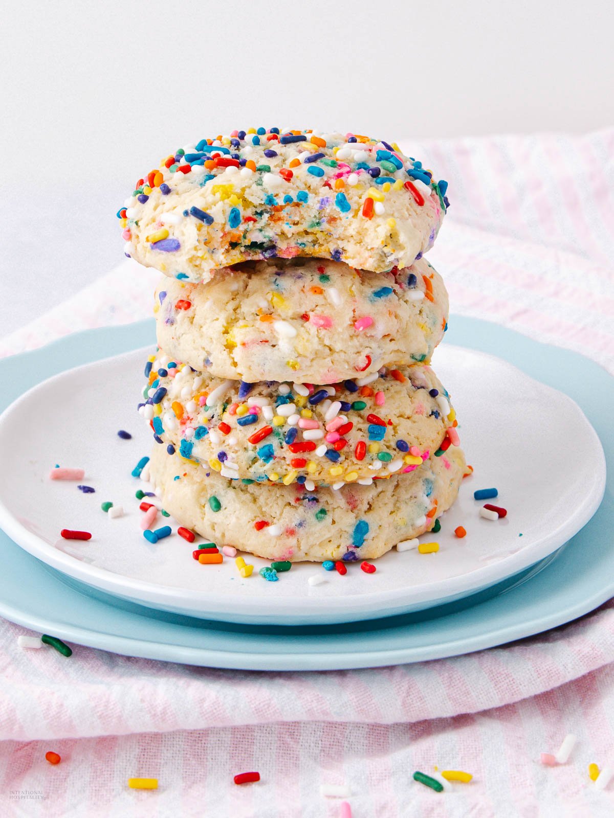 A stack of four sprinkle-covered cookies sits on a white plate with a blue rim. The top cookie has a bite taken out of it, and colorful sprinkles are scattered on the cookies and plate.