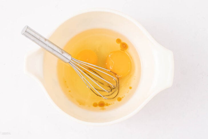 A white mixing bowl with two cracked raw eggs and a metal whisk inside, placed on a white surface.