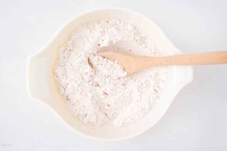 A white mixing bowl filled with flour mixture containing colorful sprinkles, being stirred with a wooden spoon, on a white surface.