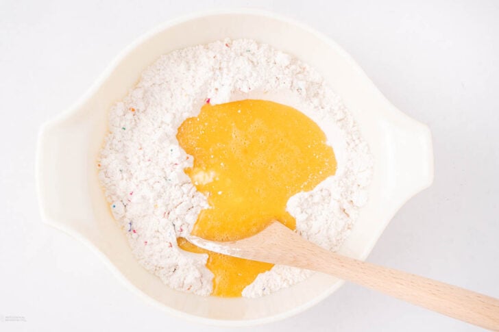 A white mixing bowl with flour and a colorful sprinkle mix on one side, and beaten eggs on the other, being stirred with a wooden spoon.