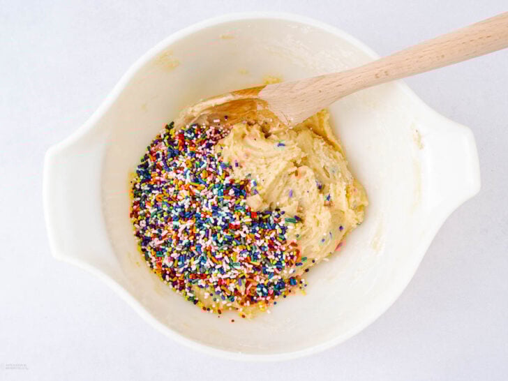 A white mixing bowl contains cookie dough and colorful rainbow sprinkles, with a wooden spoon resting inside.
