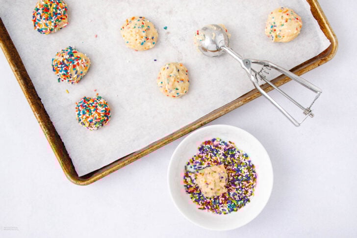 A baking tray lined with parchment paper holds several unbaked cookie dough balls with rainbow sprinkles. A cookie scoop and a small bowl of sprinkles with a dough ball inside are nearby.