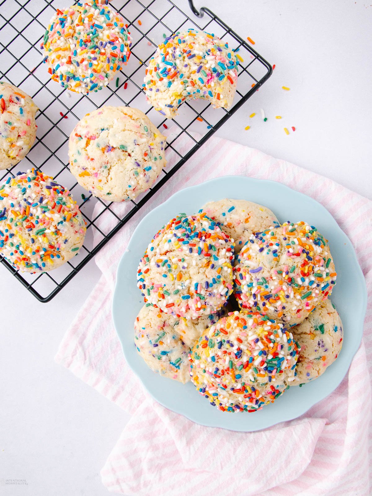 A plate and a cooling rack display several round cookies topped with colorful rainbow sprinkles, set on a light pink striped cloth against a white background.