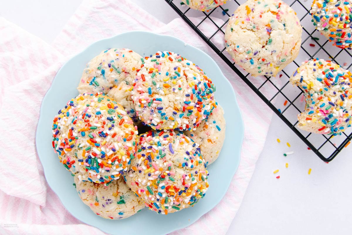 A light blue plate with a stack of cookies covered in colorful rainbow sprinkles sits on a pink striped cloth next to a cooling rack with more sprinkle cookies on a white background.