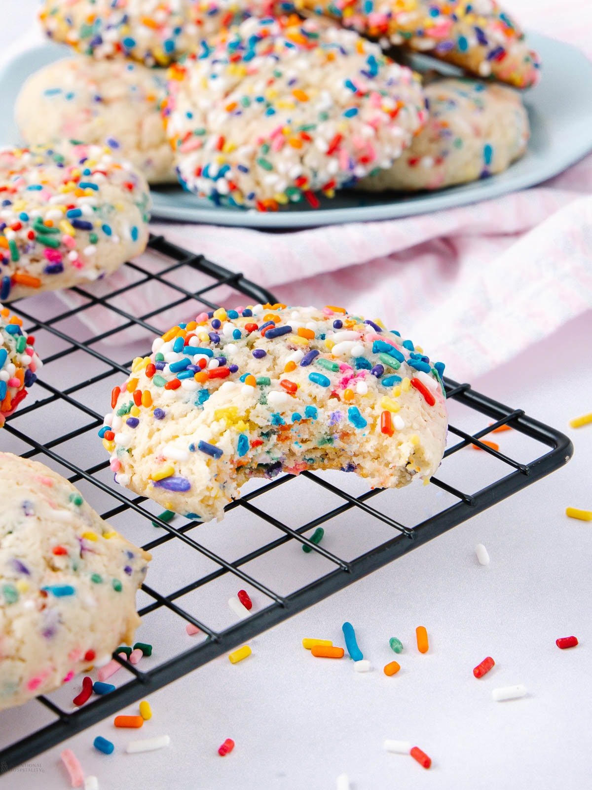 A partially eaten sprinkle cookie sits on a cooling rack with colorful sprinkles, surrounded by more whole cookies. A plate with additional cookies is visible in the background.