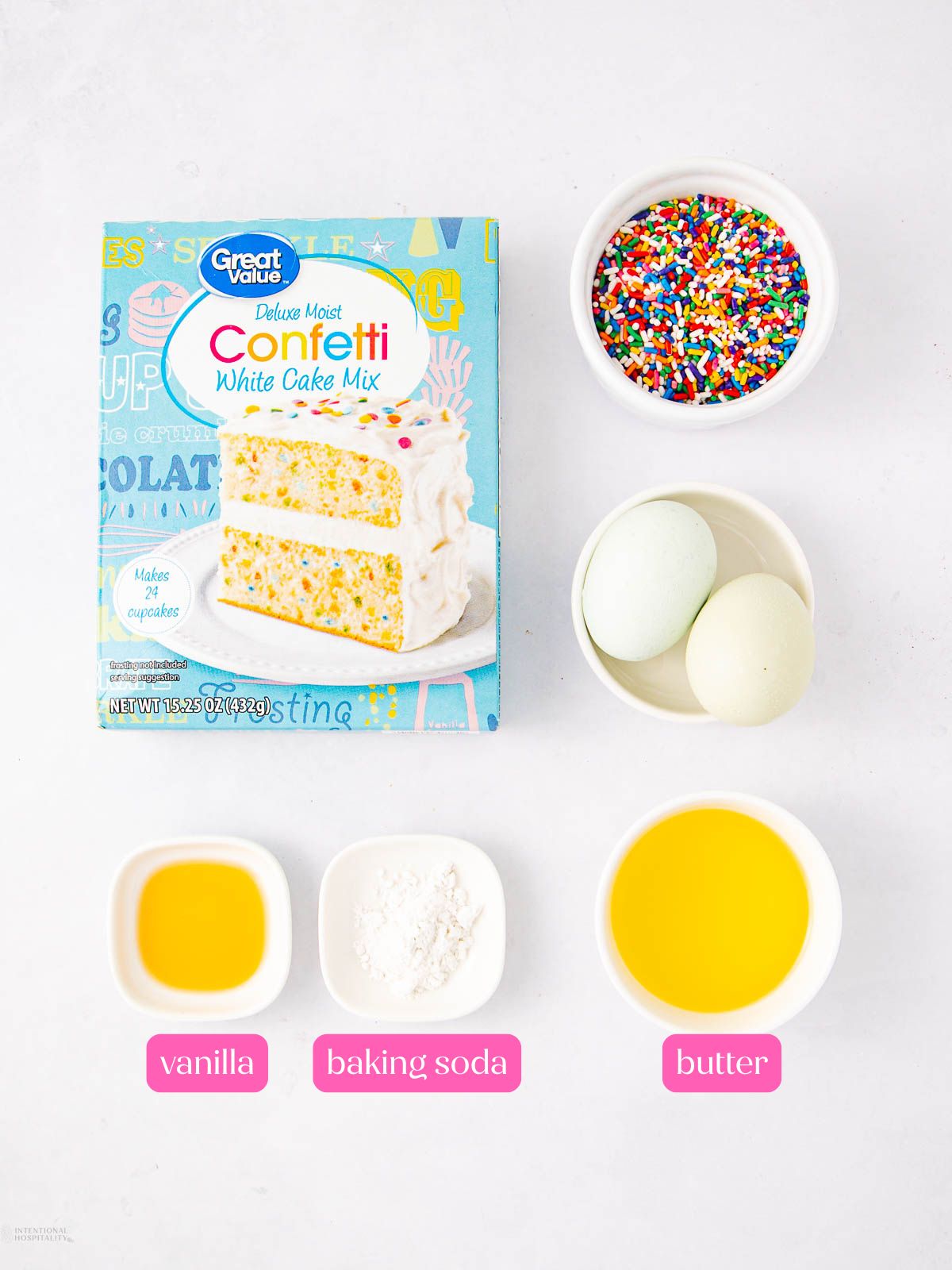 A box of confetti cake mix, a bowl of colorful sprinkles, two eggs, and small bowls labeled vanilla, baking soda, and melted butter are arranged on a white surface.