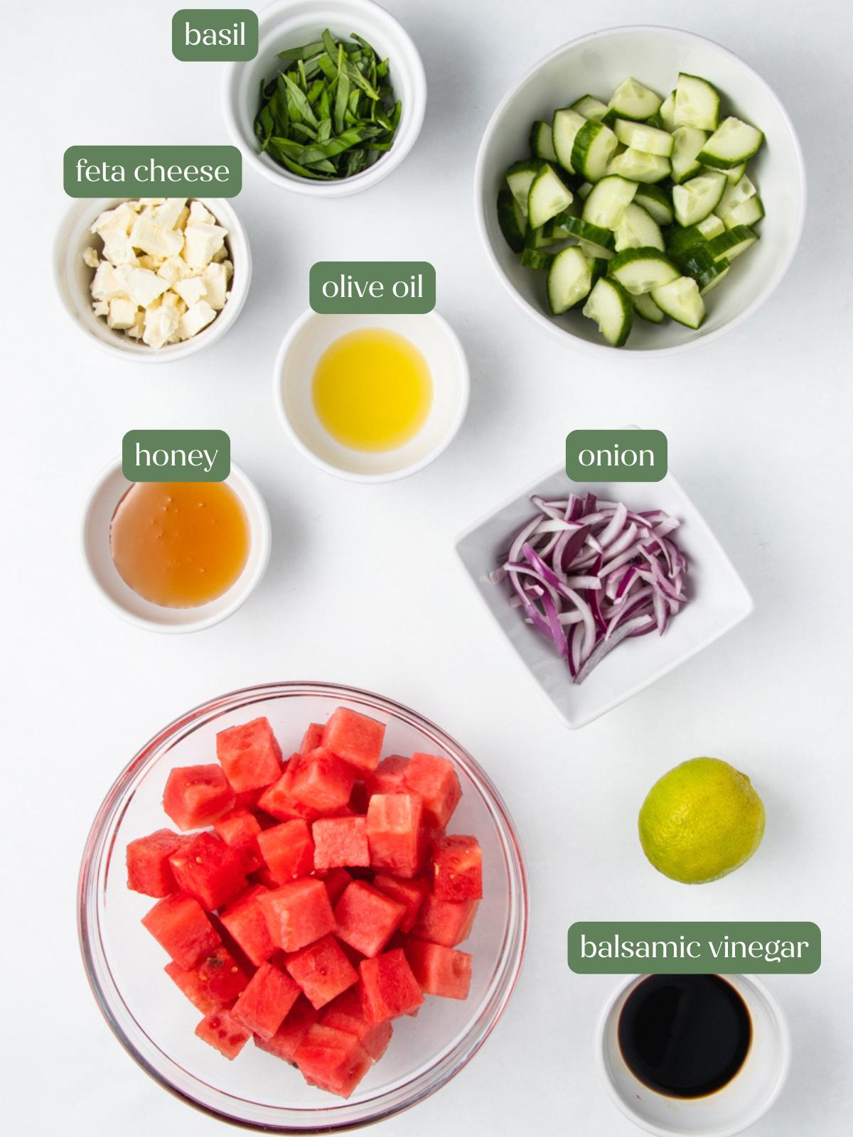 Top-down view of small bowls containing chopped basil, cubed feta cheese, olive oil, sliced cucumber, honey, sliced red onion, balsamic vinegar, a lemon, and a large bowl of cubed watermelon, all labeled.