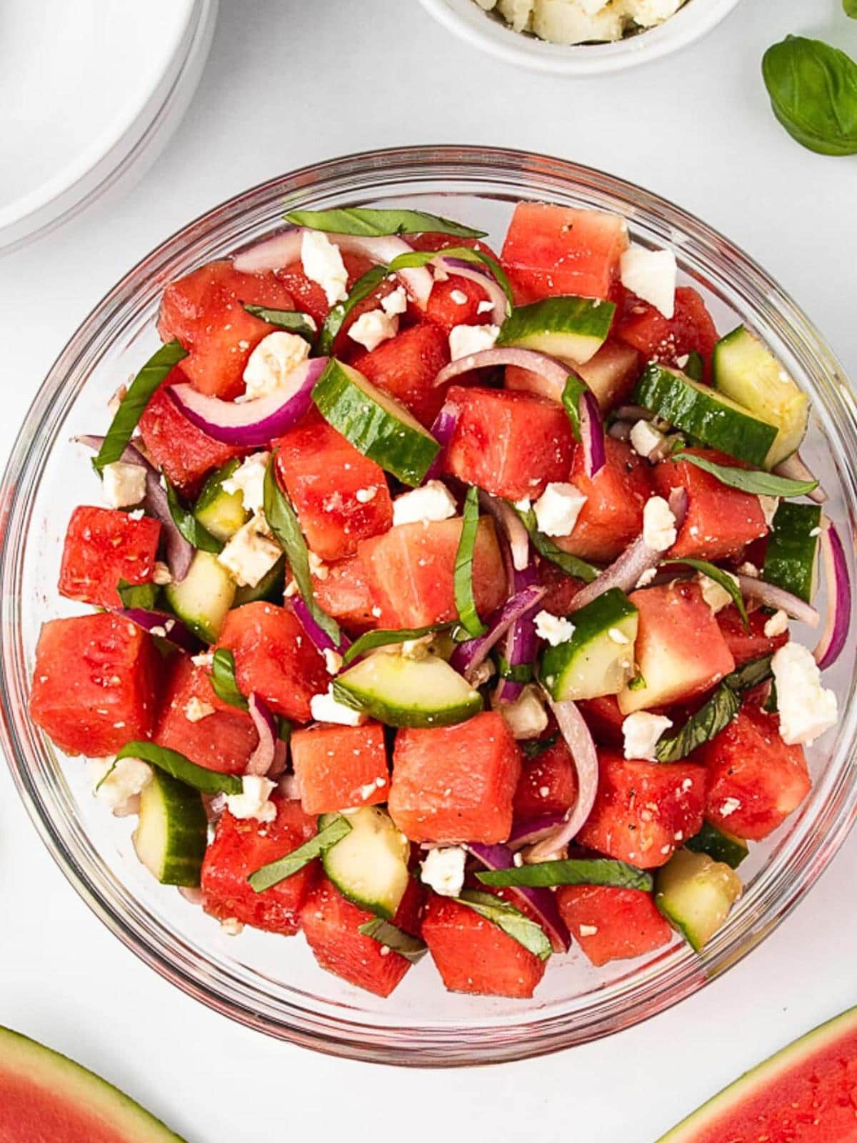 A glass bowl filled with a salad of watermelon cubes, sliced cucumbers, red onions, and crumbled feta cheese, arranged on a white surface with a partial watermelon slice nearby.