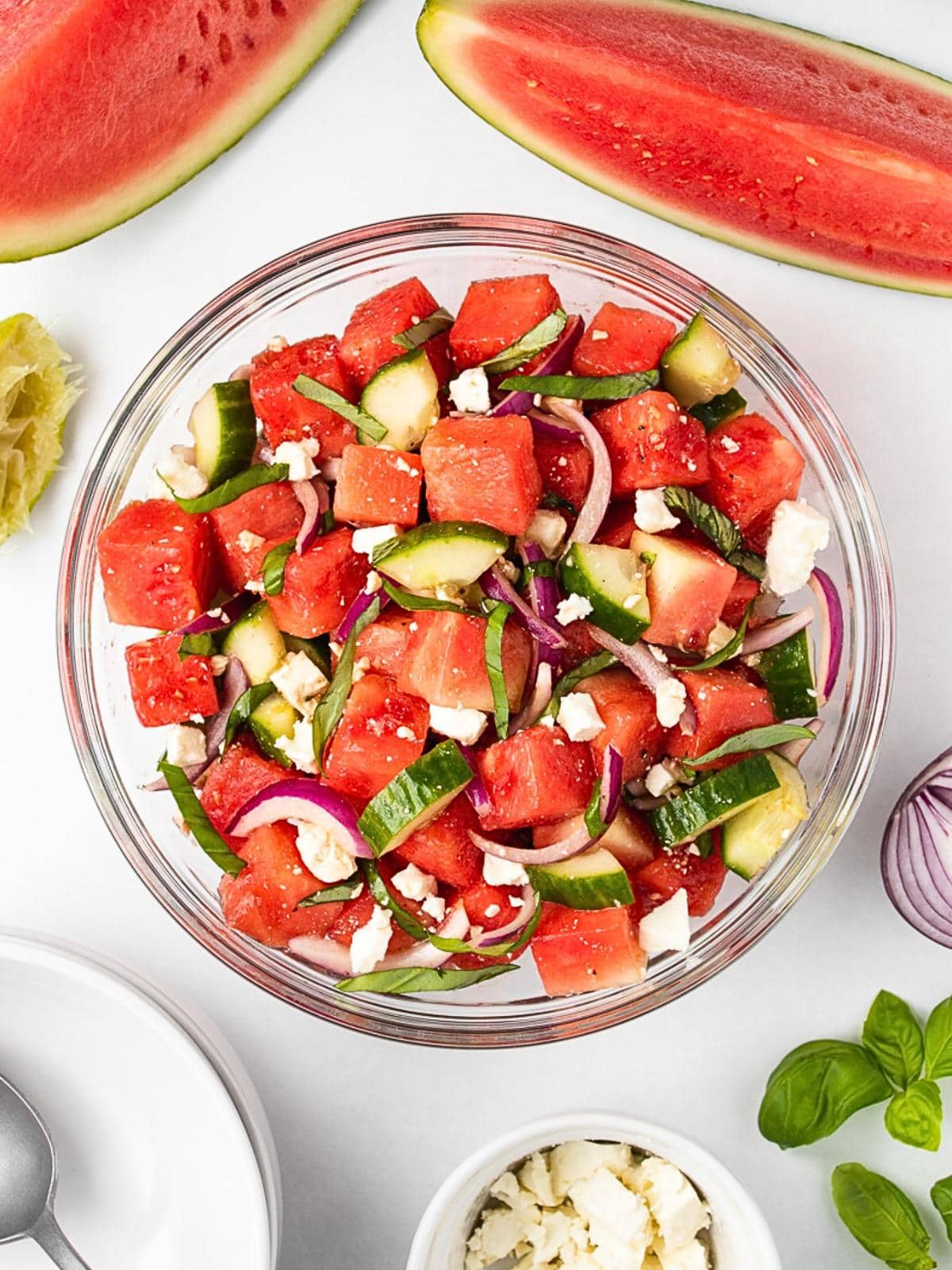 A glass bowl filled with a salad of watermelon cubes, cucumber slices, red onion, and feta cheese, surrounded by watermelon wedges, basil leaves, and a small bowl of crumbled feta on a white surface.