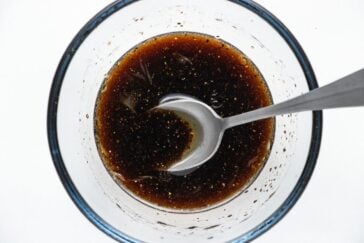 Overhead view of a glass bowl containing a dark brown liquid with visible specks, being stirred by a metal spoon. The bowl is placed on a white surface.