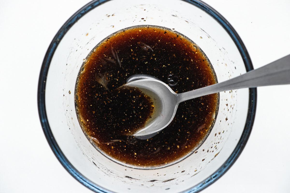 Overhead view of a glass bowl containing a dark brown liquid with visible specks, being stirred by a metal spoon. The bowl is placed on a white surface.