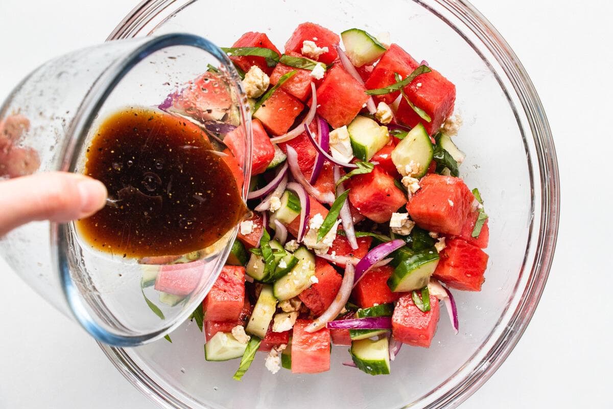 A hand pours dark vinaigrette dressing from a glass cup onto a salad with watermelon chunks, cucumber slices, red onion, crumbled cheese, and fresh herbs in a clear glass bowl.