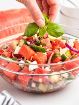 A hand places fresh basil leaves on top of a glass bowl filled with a watermelon salad containing cubes of watermelon, cucumber, red onion, and feta cheese.