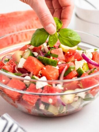 A hand places fresh basil leaves on top of a glass bowl filled with a watermelon salad containing cubes of watermelon, cucumber, red onion, and feta cheese.