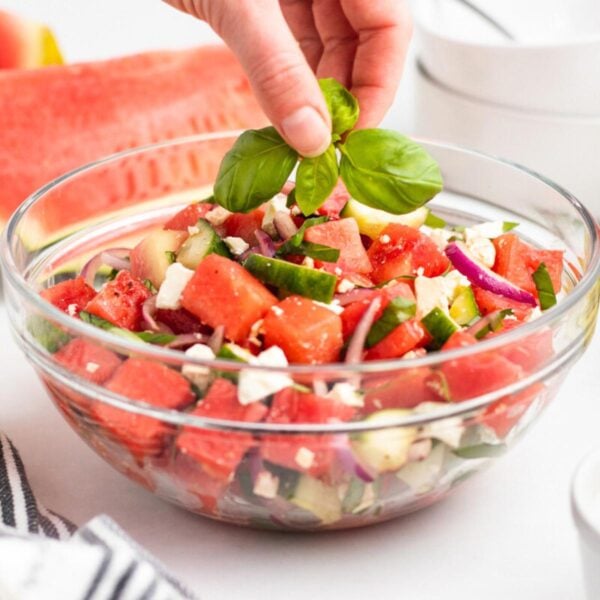 A hand places fresh basil leaves on top of a glass bowl filled with a watermelon salad containing cubes of watermelon, cucumber, red onion, and feta cheese.