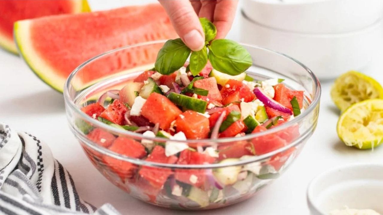 A hand places fresh basil leaves on top of a bowl of watermelon salad, which contains watermelon cubes, cucumber, red onion, and feta cheese. Sliced watermelon and a halved lime are in the background.