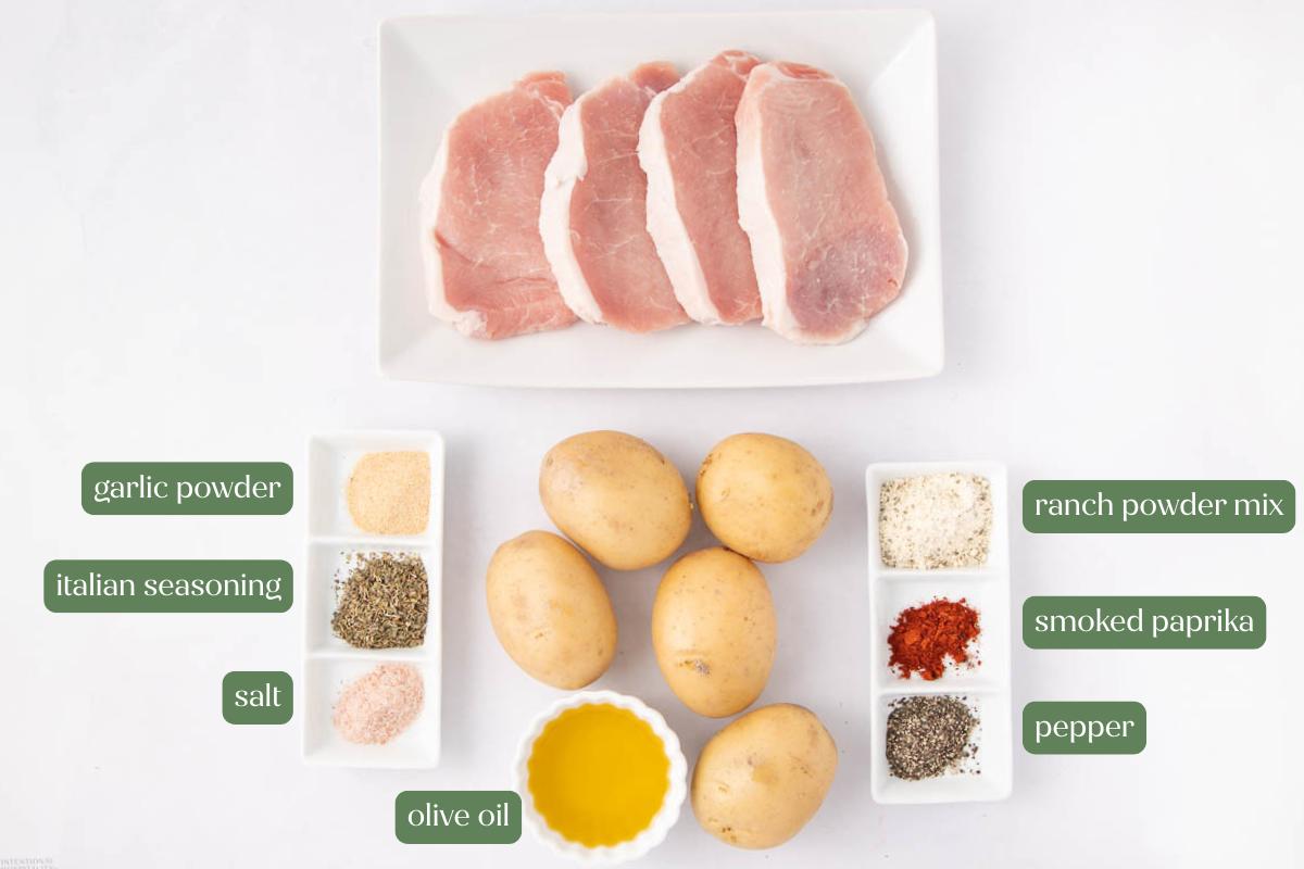 Overhead view of four raw pork chops on a plate, three whole potatoes, a dish of olive oil, and small bowls containing salt, pepper, garlic powder, Italian seasoning, ranch powder mix, and smoked paprika.