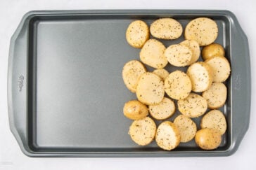 A metal baking sheet with seasoned, halved baby potatoes arranged in a cluster on one side, ready for roasting. The rest of the tray is empty.