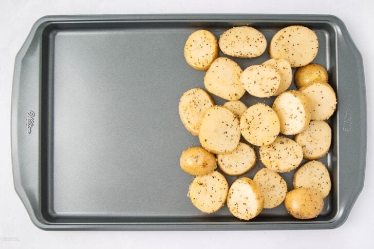 A metal baking sheet with seasoned, halved baby potatoes arranged in a cluster on one side, ready for roasting. The rest of the tray is empty.