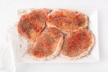 Four raw pork chops on a white plate, sprinkled heavily with a mix of dried herbs and reddish spices, likely paprika and black pepper, on a white background.