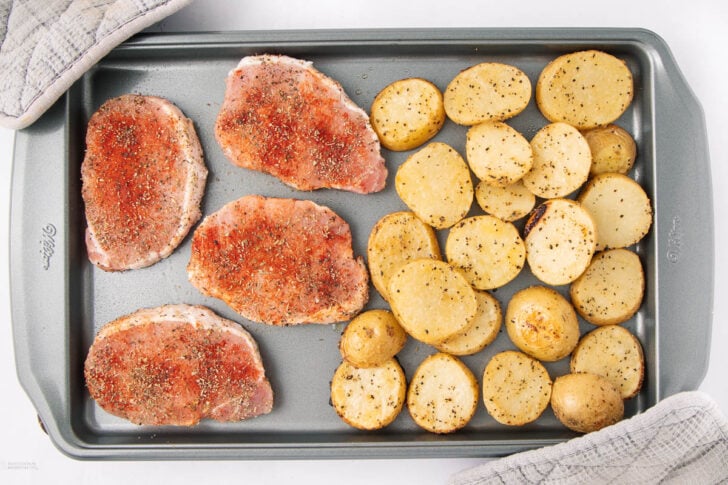 A baking sheet with four seasoned raw pork chops on the left and sliced, seasoned yellow potatoes on the right, ready to be cooked. A gray oven mitt is partially visible on the top left corner.