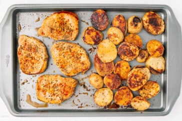A metal baking sheet with four seasoned pork chops on the left and sliced roasted potatoes on the right, both cooked to a golden brown.