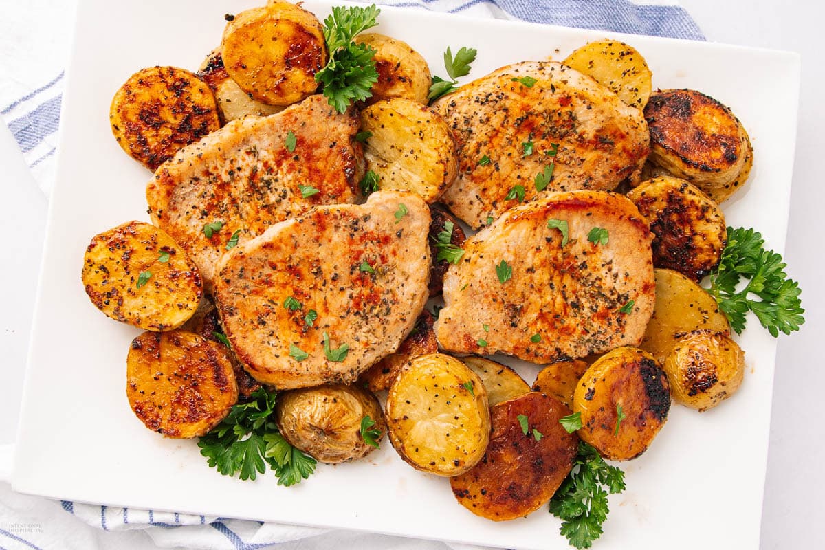 Grilled pork chops garnished with fresh parsley, served on a white plate with golden roasted potatoes. Some potatoes and herbs are scattered around for decoration.