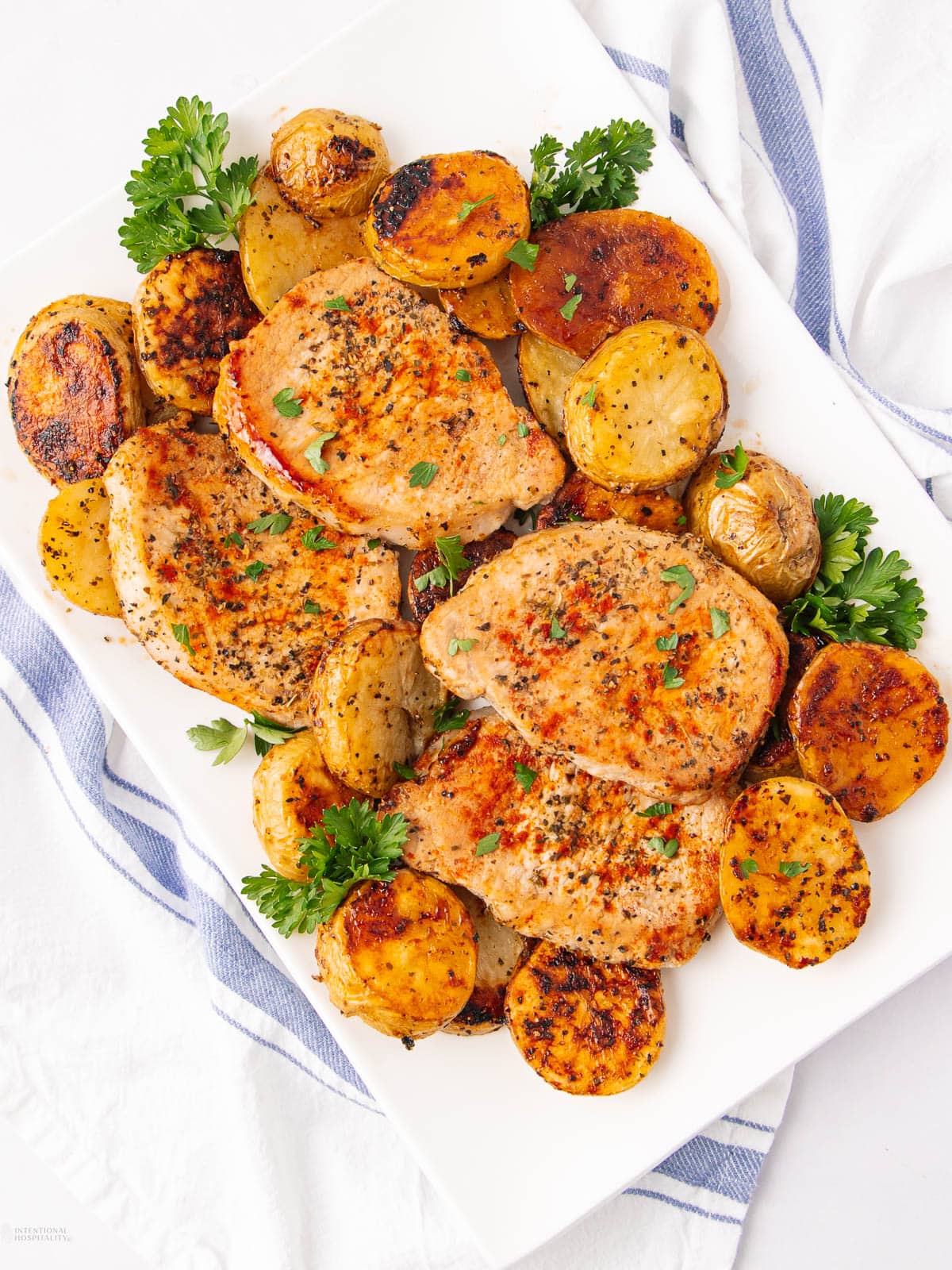 A white platter with seasoned, cooked pork chops and golden roasted potato slices, garnished with fresh parsley, on a white and blue cloth.