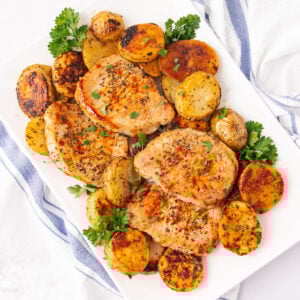 A white plate with three seasoned pork chops and roasted potatoes, garnished with fresh parsley. The plate rests on a white surface with a blue and white striped cloth nearby.