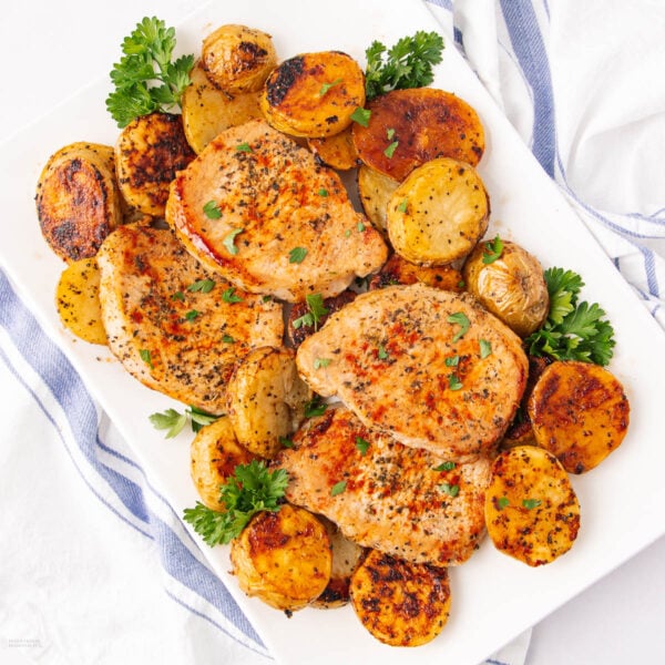 A white plate with three seasoned pork chops and roasted potatoes, garnished with fresh parsley. The plate rests on a white surface with a blue and white striped cloth nearby.