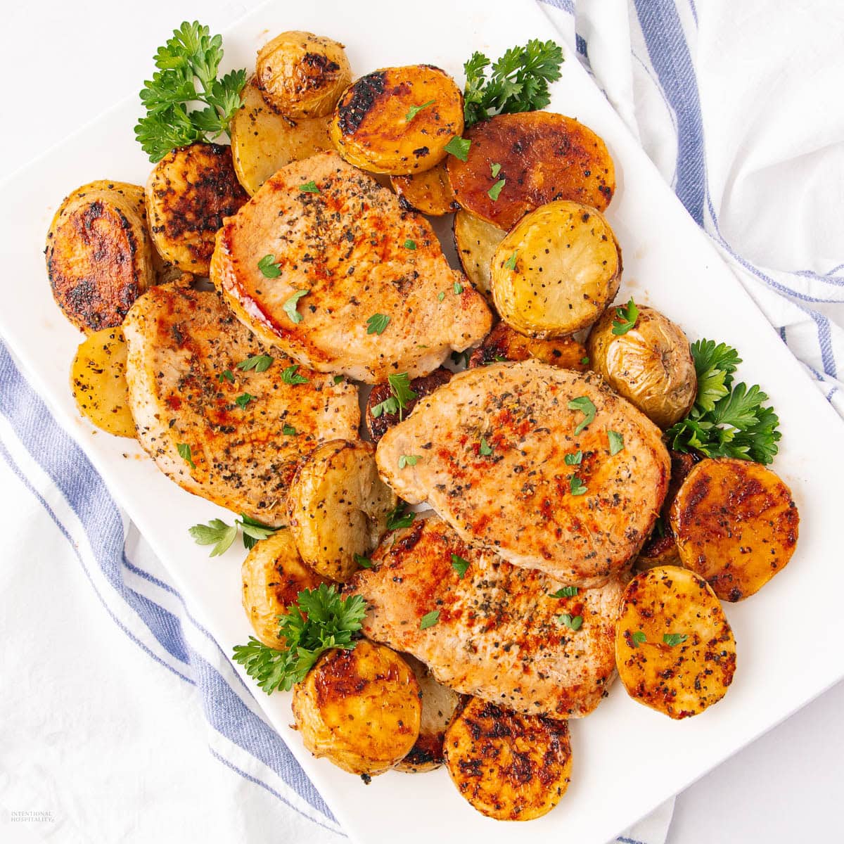 A white plate with three seasoned pork chops and roasted potatoes, garnished with fresh parsley. The plate rests on a white surface with a blue and white striped cloth nearby.