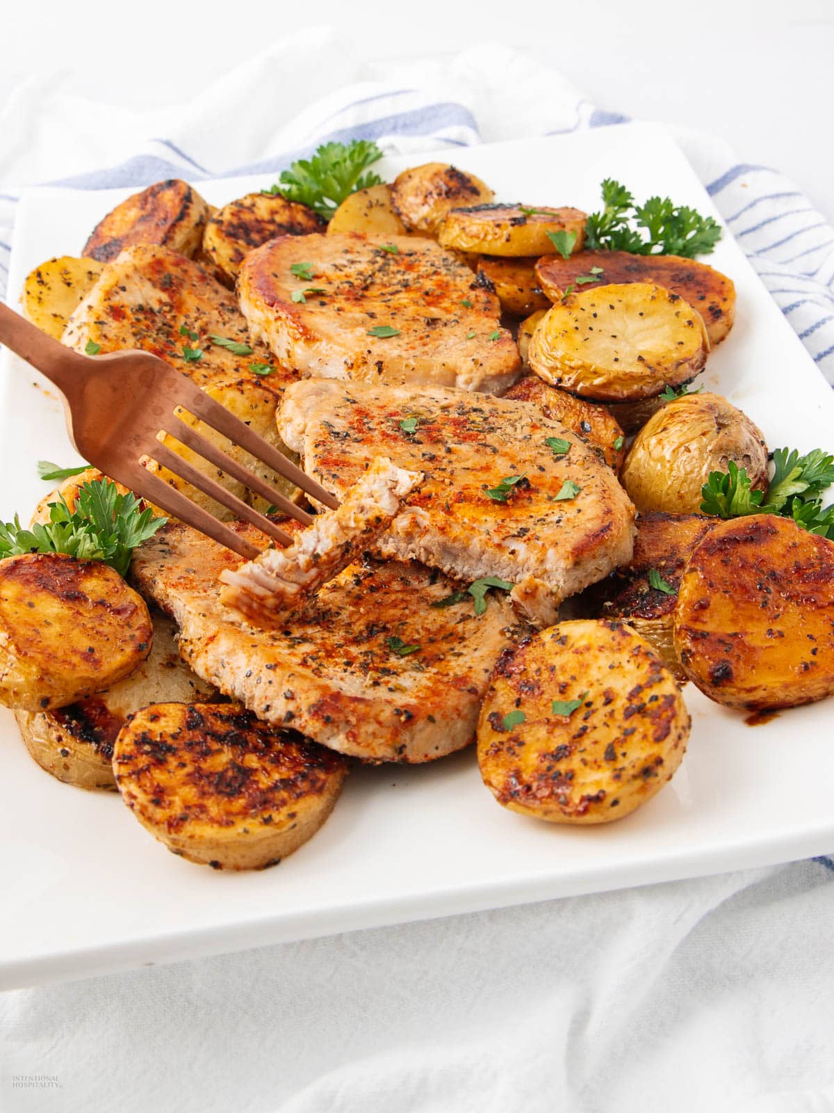 A white rectangular plate with golden-brown pork chops and roasted potato slices, garnished with parsley. A fork holds a piece of pork chop, showing its tender, juicy texture.