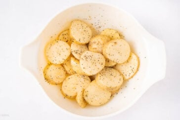 Sliced potatoes in a white bowl, seasoned with black pepper and spices, ready for cooking. The bowl is placed on a white surface.