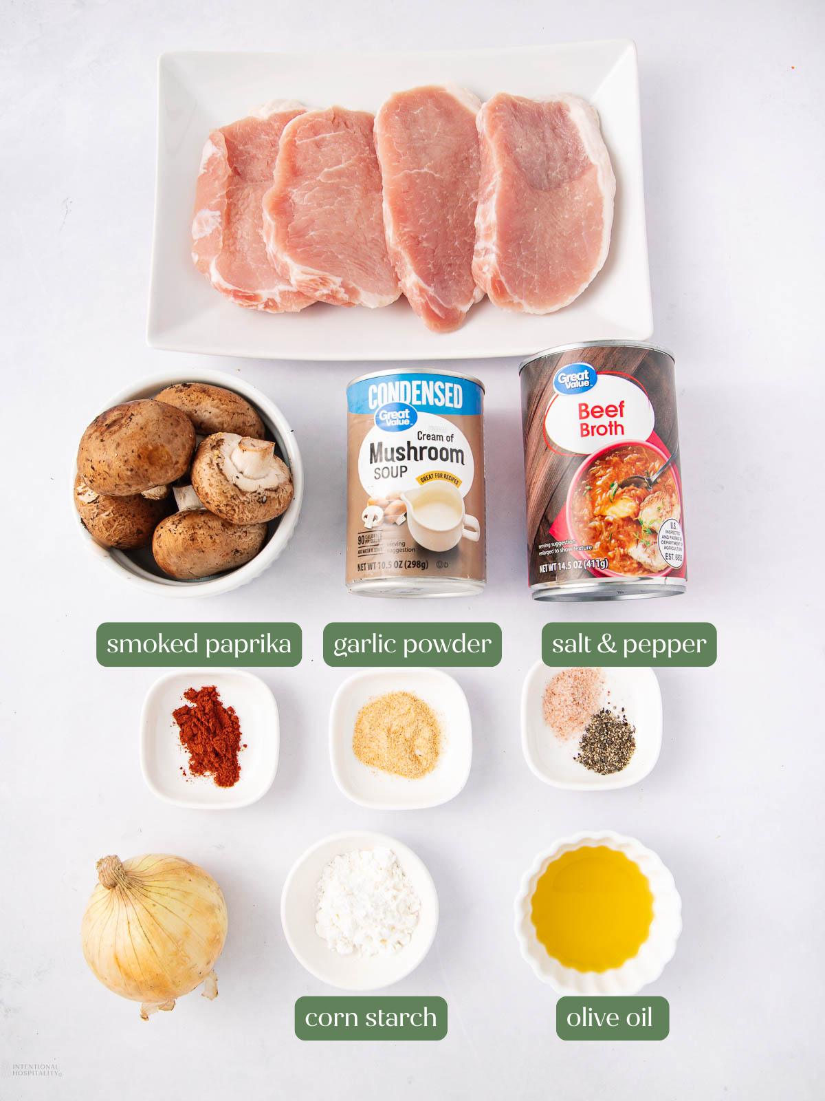 Top-down view of raw pork chops, whole mushrooms, a can of cream of mushroom soup, a carton of beef broth, seasonings (smoked paprika, garlic powder, salt and pepper), cornstarch, an onion, and a bowl of olive oil.