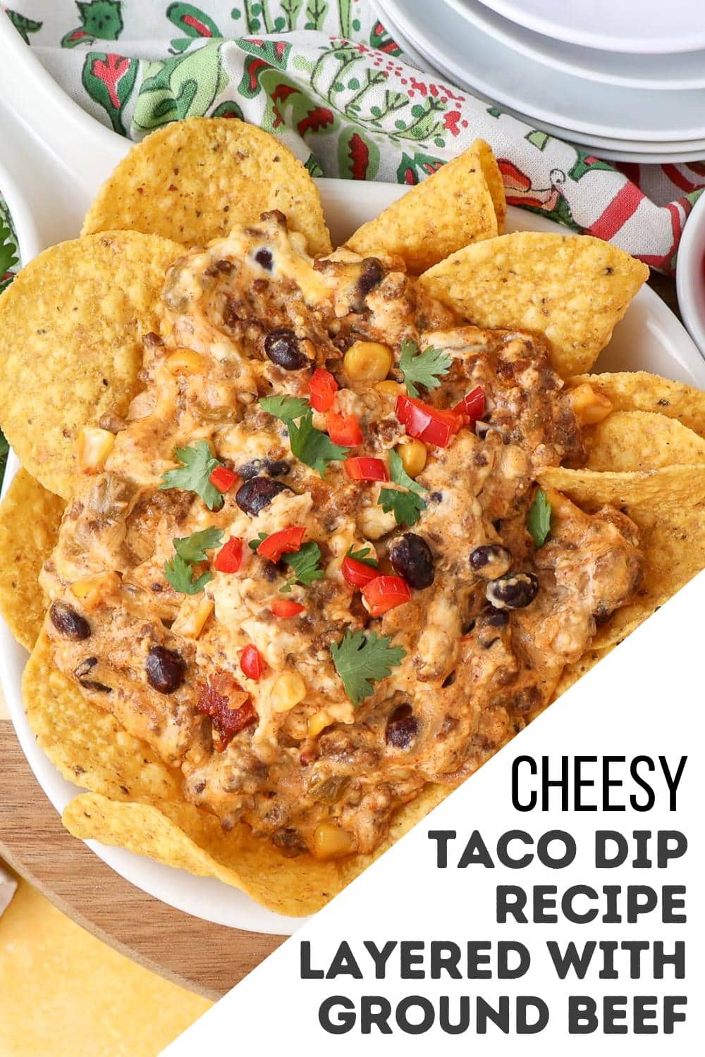 A plate of tortilla chips surrounds a cheesy taco dip topped with ground beef, black beans, corn, melted cheese, chopped red peppers, and cilantro. The text on the image reads: "Cheesy taco dip recipe layered with ground beef.
